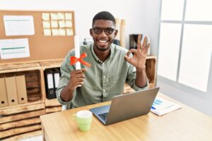 Young african american student man holding graduate degree diploma sitting on the table doing ok sign with fingers, smiling friendly gesturing excellent symbol